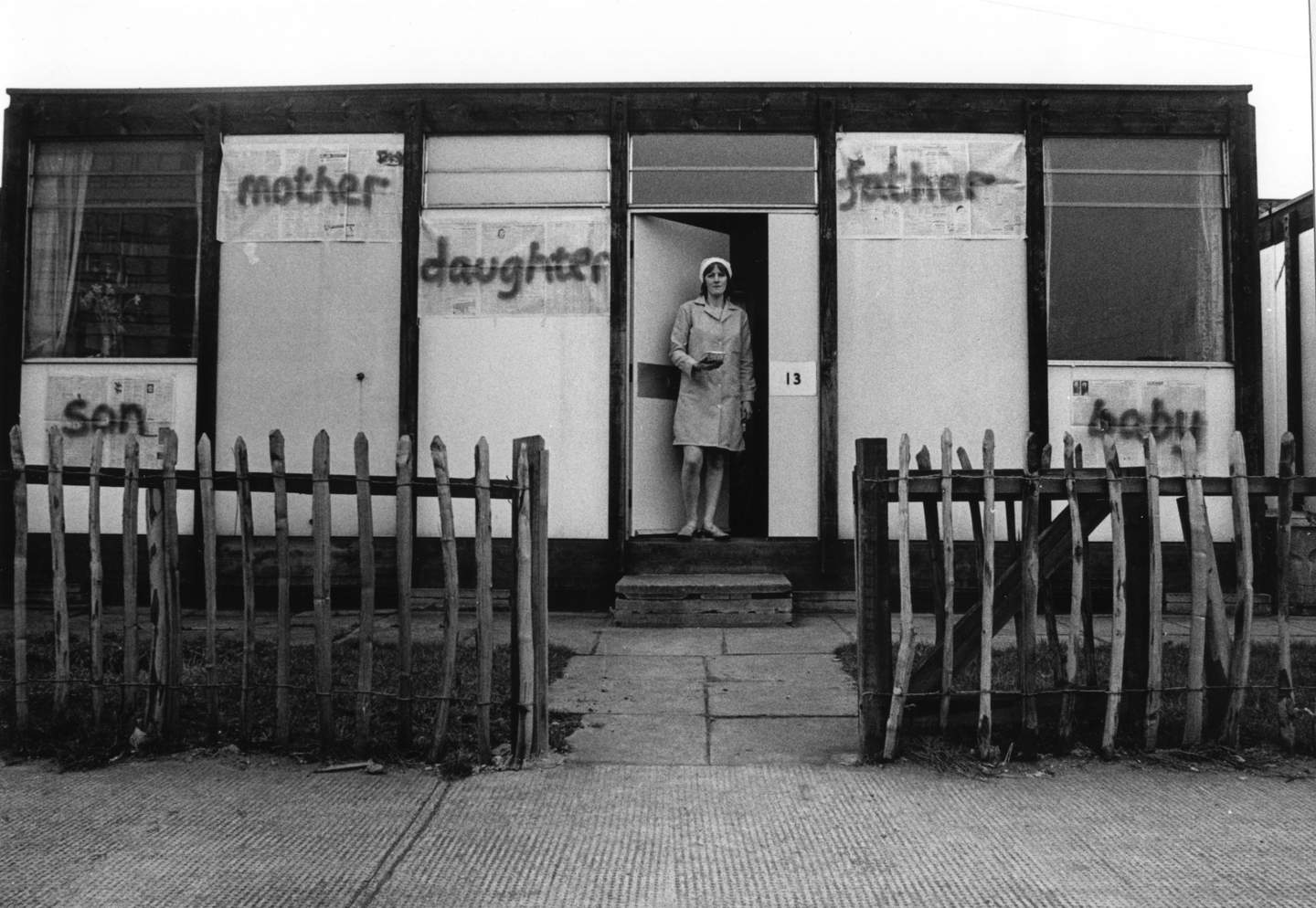 Black and white photograph from the 70s of a woman standing on the doorstep of a prefab building. Newspaper sheets with 