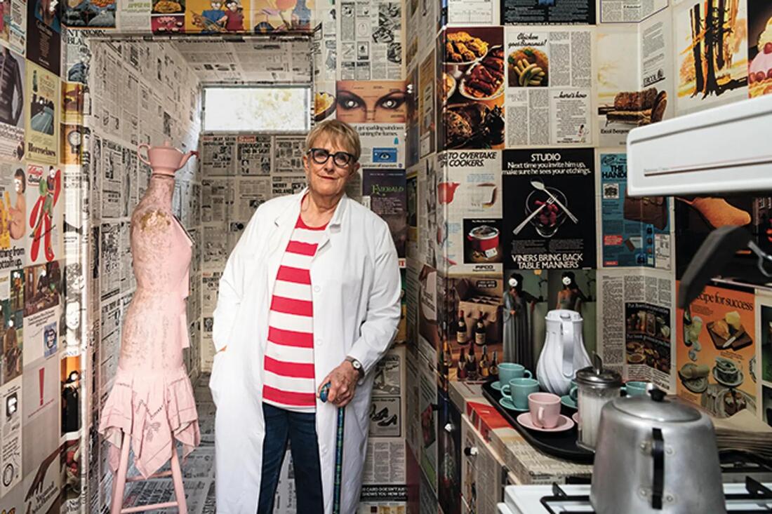 A woman smiling and wearing a white lab coat stands in a room whose walls are plastered in newspaper sheets, things to make tea are seen in the foreground