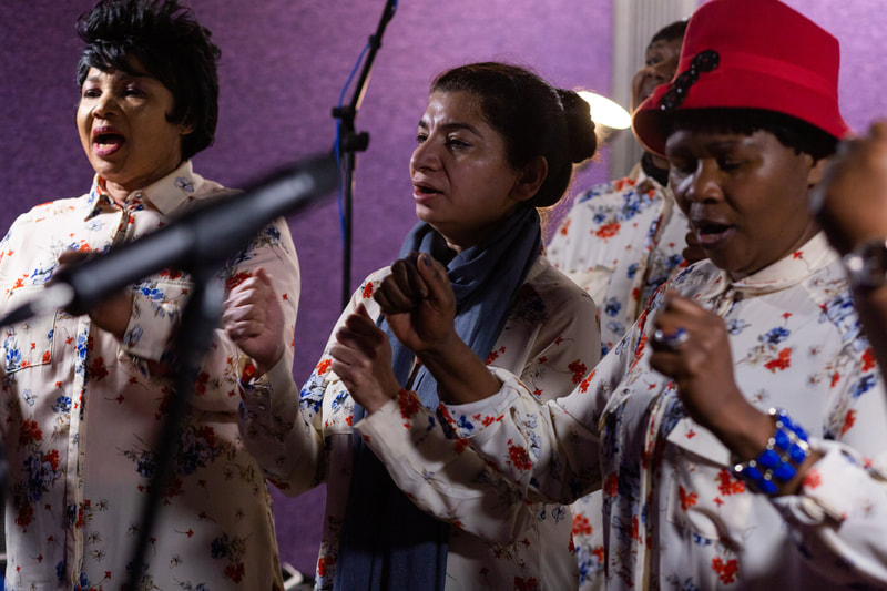 three women wearing matching shirts sing and clap in front of professional microphones